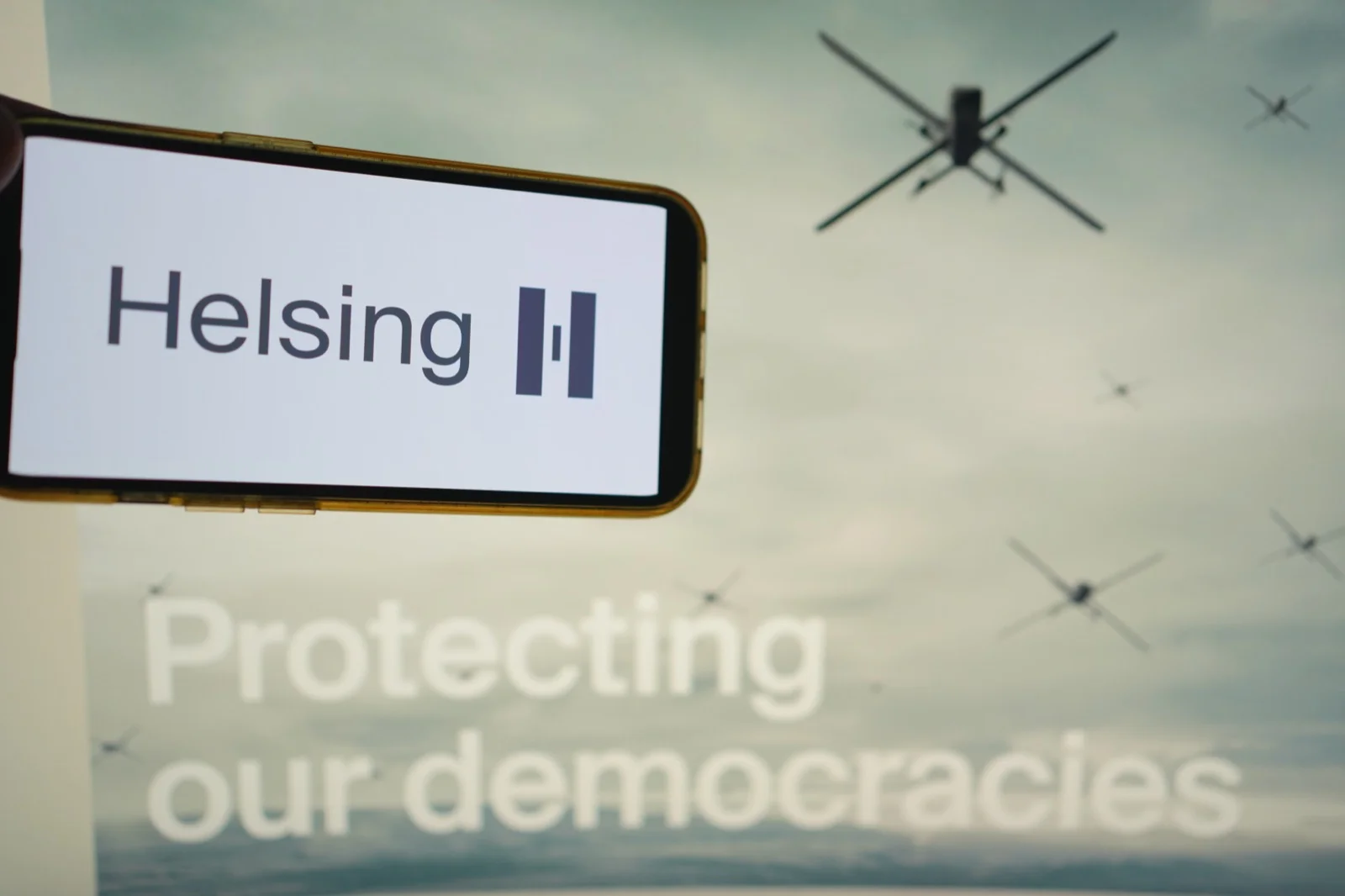 Phone screen displaying the Helsing logo against a backdrop of military drones and the company's "Protecting our democracies" tagline. The Munich-based defence AI company is the largest private AI company in Europe, valued at $14.1 billion as of June 2025.